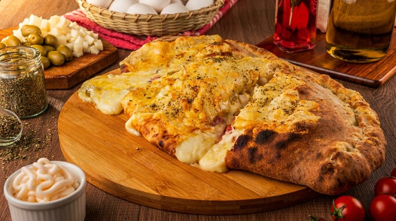 Cheesy calzone slices on a wooden board, surrounded by olives, spices, and sauces.