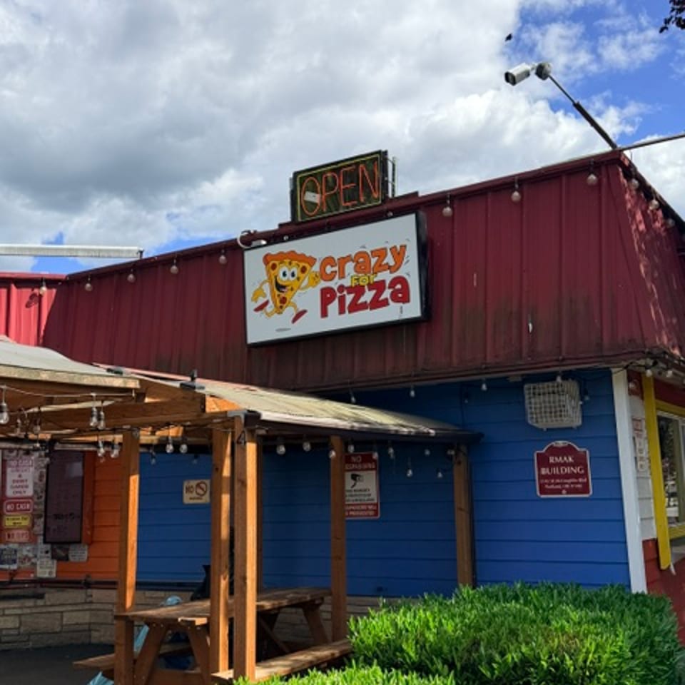 Crazy For Pizza storefront with outdoor seating, signage, and green landscaping.