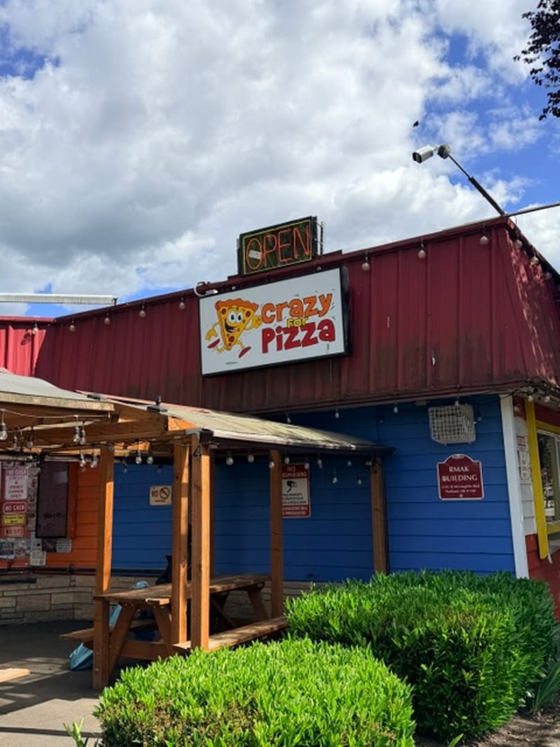 Crazy For Pizza storefront with outdoor seating, signage, and green landscaping.