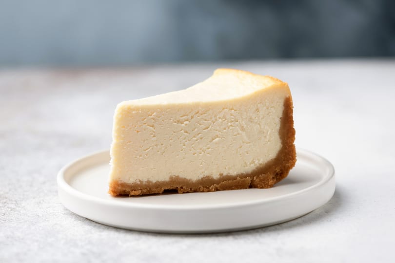 A slice of classic cheesecake with a graham cracker crust on a white plate.