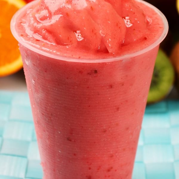 A vibrant red strawberry smoothie in a clear plastic cup, surrounded by fresh fruit.