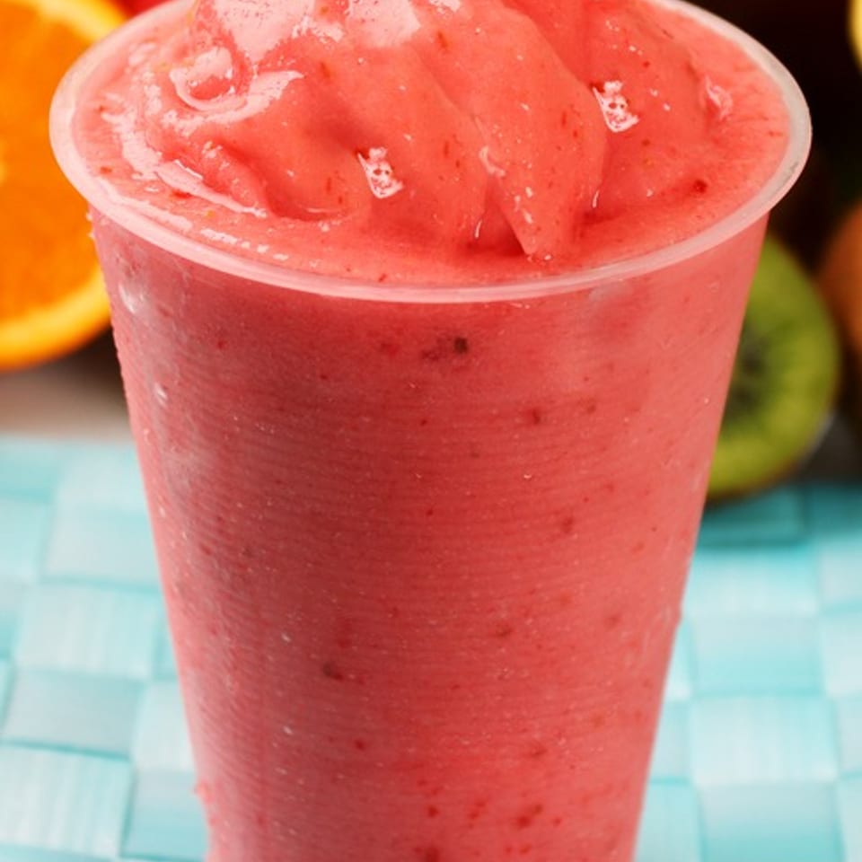 A vibrant red strawberry smoothie in a clear plastic cup, surrounded by fresh fruit.